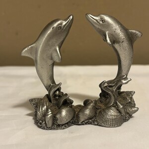 May include: A silver-colored dolphin figurine with two dolphins leaping towards each other. The dolphins are mounted on a base with seashells. The figurine is made of metal and has a detailed design.