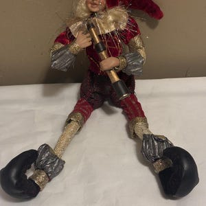 May include: A decorative elf figurine with a red velvet hat and suit, gold accents, and black shoes. The elf holds a gold and black telescope-like object. The figurine has blonde hair and a smiling face. The elf is posed with legs spread.