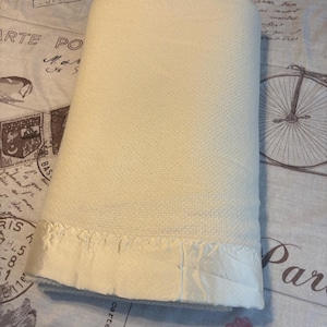 May include: A folded off-white thermal blanket featuring a textured waffle-knit pattern and a smooth, shiny satin border trim along the bottom edge. The blanket is rectangular in shape and displays a consistent light cream color throughout the fabric.