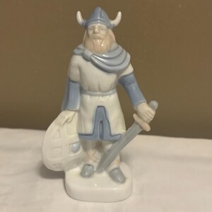 May include: A porcelain figurine of a Viking warrior wearing a horned helmet, a tunic, and a cape. The figure is colored in shades of white and light blue, holding a sword in one hand and resting the other hand on a round shield at the side.