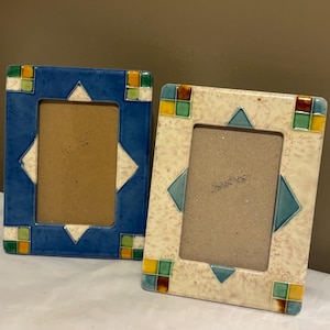 May include: Two ceramic picture frames. One is blue with white and green accents, the other is off-white with teal and brown accents. Both have a geometric design with a rectangular opening for a photo.