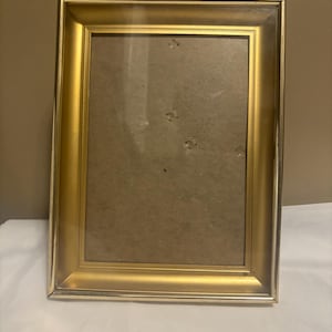 May include: A rectangular gold-colored picture frame with a glass front. The frame has a glossy finish and a simple, elegant design. The interior is a neutral tan color, ready for a photo or artwork. The frame is suitable for displaying photos or artwork.