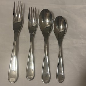 May include: Four pieces of stainless steel silverware are arranged on a white surface. The set includes two forks and two spoons. The forks have four tines, and the spoons have oval bowls.