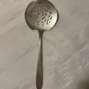May include: A silver-colored serving spoon with a round, pierced bowl featuring an ornate floral design. The handle is long and slender, with decorative etching. This vintage utensil is likely for serving food.