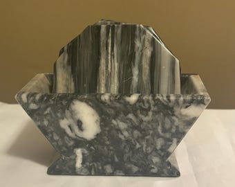 May include: A set of marble coasters in a holder. The coasters and holder are gray and white with a marbled pattern. The holder has a geometric shape with a flared base.