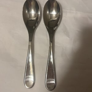 May include: Two shiny, silver spoons with oval bowls and teardrop-shaped handles. The spoons are placed side-by-side on a white surface, reflecting light. The spoons appear to be made of stainless steel and are likely for eating.