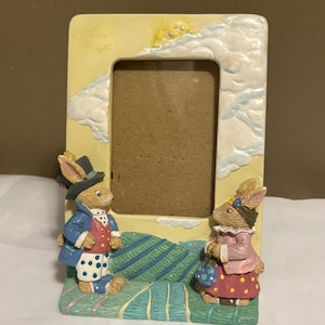 May include: A decorative picture frame featuring two rabbit figurines. The frame has a yellow background with a sun and clouds. One rabbit wears a blue coat and top hat, the other a pink dress and bow.