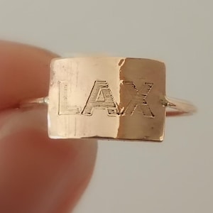 May include: A gold-colored ring with a rectangular face. The word "LAX" is engraved on the face of the ring. The ring is held up by a hand against a neutral background.