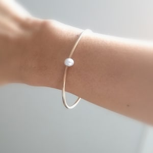 May include: A simple gold bangle bracelet with a single white pearl accent.