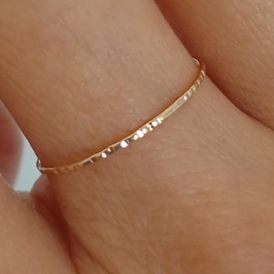 Puede incluir: Un delicado anillo de oro con una superficie texturizada. El anillo es delgado y se encuentra en un dedo, mostrando su diseño simple pero elegante. El cálido color dorado del anillo contrasta con el tono de la piel, resaltando sus sutiles detalles.