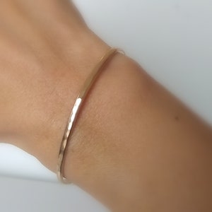 Pode incluir: Um bracelete simples de ouro martelado. O bracelete é fino e delicado, e é mostrado no pulso de uma pessoa.