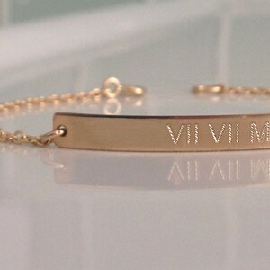 May include: A gold-colored bracelet with a rectangular bar and delicate chain. The bar is engraved with Roman numerals. The bracelet is displayed on a reflective surface, highlighting its polished finish. A simple and elegant piece of jewelry.