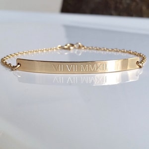 May include: A gold-colored bracelet with a rectangular bar engraved with Roman numerals VII VII MMXII. The bracelet has a delicate chain and clasp. The jewelry is displayed on a white surface.