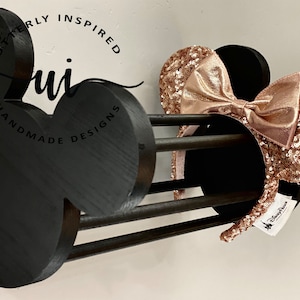 May include: Black wooden Mickey Mouse-shaped headband holder with a rose gold sequined headband with a bow. The holder has the text "Utterly Inspired" and "Handmade Designs" on it.