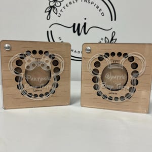 May include: Two wooden baby tooth keepsake boxes. Each box has a clear acrylic overlay in the shape of Mickey Mouse ears. The text "Upper" and "Lower" are visible. One box says "Paxton's Baby Teeth" and the other says "Watt's Baby Teeth".