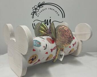 May include: A light-colored wooden display stand shaped like a mouse head, holding a cylindrical pillow. The pillow is covered in a white fabric with a pattern of cartoon characters and red bows. A pair of sequined mouse ears with a bow are on top.