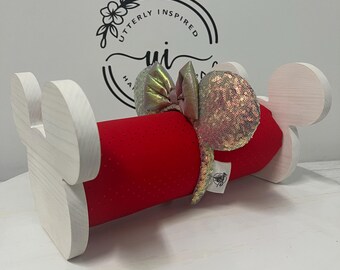 May include: A red cylindrical holder with white wooden Mickey Mouse ear accents, holding a sequined bow headband. The headband has a pink bow and iridescent sequin ears. The holder is branded with the text "Utterly Inspired".