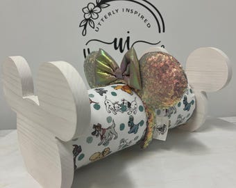 May include: A white wooden Mickey Mouse ear holder with a Dalmatian print headband. The headband has a sequined ear and a bow with iridescent and pink hues. The holder has the text "Utterly Inspired" on the back.