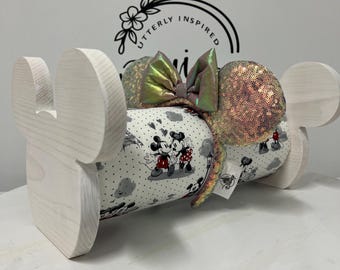 May include: A wooden Mickey Mouse ear holder in a light wood finish. The holder displays a sequined ear headband with a large iridescent bow. The headband rests on a fabric-covered cylinder featuring Mickey and Minnie Mouse.