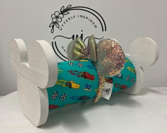 May include: A white wooden Mickey Mouse ear holder with a teal fabric cylinder featuring race cars and checkered flags. The ears are iridescent sequins with a matching bow.