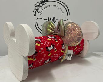 May include: A wooden Mickey Mouse ear holder with a red fabric center featuring a Mickey and Minnie Mouse pattern. The ears are embellished with iridescent sequins and a matching bow. The holder is white.