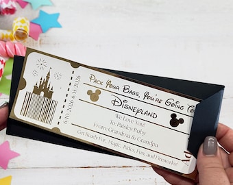 May include: A rectangular paper ticket with gold foil accents on a black cardstock holder. The ticket features a castle graphic, a barcode, and text reading: Pack Your Bags, You're Going To Disneyland, 6/10/2026-6/15/2026, We Love You! To: Paisley Ruby, From: Grandma & Grandpa, Get Ready For, Magic, Rides, Fun, and Fireworks!