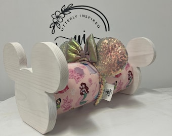 May include: A white wooden Mickey Mouse ear holder with a pink fabric roll featuring Disney princesses. The holder has a bow and sequined ears. The text "Utterly Inspired" is visible in the background.