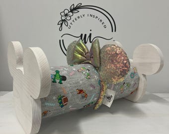 May include: A light-colored wooden display stand with a cylindrical center wrapped in gray fabric featuring cartoon characters. The stand has large, round ears and a bow with iridescent sequins. The fabric has the word "GOAL" printed on it.