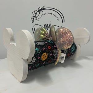 May include: A white wooden display stand shaped like Mickey Mouse ears holds a pair of sequined mouse ears. The ears are iridescent with a black fabric band featuring colorful sugar skull designs. The text "Utterly Inspired" is visible in the background.