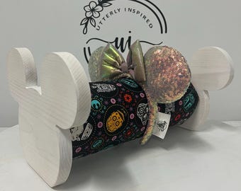 May include: A white wooden display stand shaped like Mickey Mouse ears holds a pair of sequined mouse ears. The ears are iridescent with a black fabric band featuring colorful sugar skull designs. The text "Utterly Inspired" is visible in the background.