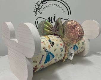 May include: A white wooden Mickey Mouse ear holder with a decorative Disney ear headband. The headband features a colorful print with Disney characters and iridescent sequins. The holder has a decorative floral logo in the background.