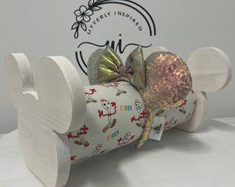 May include: A white wooden Mickey Mouse ear holder with a Forrky-themed headband. The headband has sequined ears and a bow with iridescent and pink accents. The holder is made of light-colored wood.