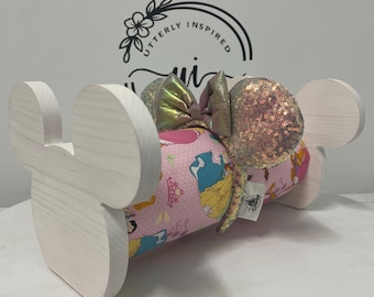 May include: A white wooden Mickey Mouse ear holder with a pink princess-themed bow and sequined ears. The bow has a gold iridescent sheen. The holder is designed to display headbands and hair accessories.