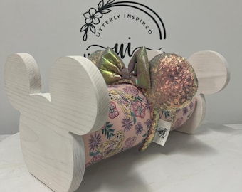 May include: A light-colored wooden Mickey Mouse ear holder with a pink floral-patterned pillow featuring a Disney princess design. The pillow holds a pair of sequined ears with a matching iridescent bow. The ears are rose gold in color.