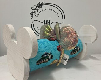 May include: A white wooden Mickey Mouse ear holder with a Cars-themed headband. The headband features a teal fabric with a map design and images of the Cars characters. The ears are embellished with iridescent sequins and a bow.