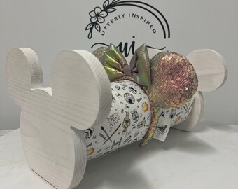 May include: A wooden Mickey Mouse ear holder in a natural finish. The holder displays a pair of sequined ears and a bow. The ears are gold and the bow is iridescent. The holder also displays a pair of ears with a white and black pattern.
