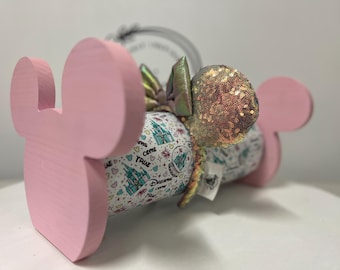 May include: A pink wooden Mickey Mouse ear holder with a pair of sequined ears and a bow. The ears are gold and the bow is iridescent. The holder is painted light pink and has a cylindrical shape.