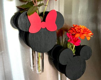 May include: Two black wooden wall-mounted planters shaped like mouse heads, one featuring a red bow. Each planter includes a clear glass test tube holding fresh flowers, including red roses and orange zinnias, attached to a metallic surface.