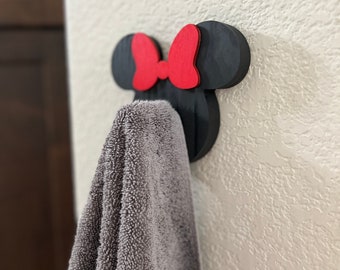 May include: Black wooden wall hook shaped like a cartoon mouse head with a red bow. A grey towel is hanging on the hook.