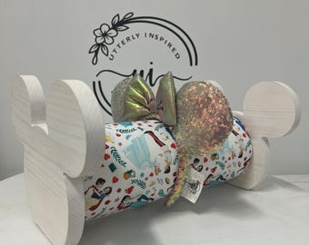 May include: A white wooden Mickey Mouse ear holder with a cylindrical center wrapped in fabric featuring Disney princess characters. The holder displays a pair of sequined mouse ears with a bow.
