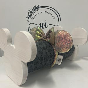 May include: A wooden Mickey Mouse ear holder in a light color. The holder supports a headband with sequined ears and a bow. The headband has a gray pattern with a Haunted Mansion theme. The bow has a shimmering, iridescent finish.
