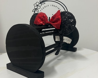 May include: Black sequined Minnie Mouse ears with a red sequined bow, displayed on a black wooden stand. The stand has a circular design with the text "Utterly Inspired" in a floral design in the background.