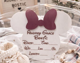 May include: A white, Mickey Mouse-shaped sign with a burgundy bow, personalized with the name "Audrey Grace Bostic." The sign includes spaces for date, time, weight (lbs, oz), and length.  A pair of cream-colored baby booties and a wooden bead bracelet are also visible.
