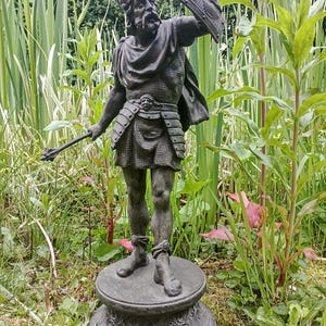 May include: A bronze statue of a warrior figure, holding a shield and a weapon. The statue is detailed with armor and a helmet, standing on a decorative base. The statue is set outdoors among green plants.