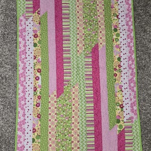 May include: A colorful quilt with a patchwork design. The quilt features vertical stripes in shades of pink, green, and yellow, with floral and geometric patterns. The edges are finished with a green border.