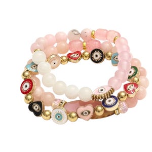 May include: Three stacked bracelets with pink and white beads. The bracelets have gold accents and charms with eye and heart designs in red, black, and blue. A stylish accessory for any outfit.