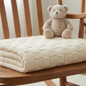 Op de afbeelding: Een lichtbruine teddybeer zit op een gevouwen crèmekleurige gebreide deken op een houten schommelstoel. De stoel heeft een gebogen rugleuning en armleuningen. Op de achtergrond is een wieg te zien.