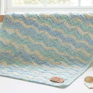 May include: A handmade, blue, green, and cream-coloured crochet baby blanket with a wave pattern. The blanket is draped over a cot, with a wooden toy and a small stack of cream-coloured cloths nearby. A leather tag reads "Cozy Cuddles."
