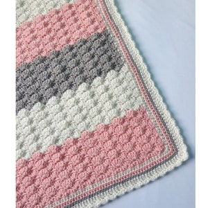 May include: A crocheted baby blanket with a textured stitch pattern in pink, white, and gray colors. The blanket has a crocheted border in pink, white, and gray.