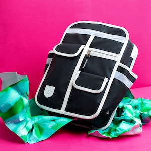 May include: Black backpack with white trim, featuring multiple pockets and a zipper closure. The bag has reflective stripes and a white emblem. The background is a vibrant pink, with a splash of green and blue wrapping paper.